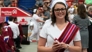 Métis nursing graduate stands holding their diploma while wearing Ceinture Fléchée
