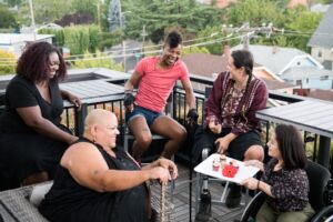 A South Asian person in a wheelchair brings mini cupcakes out on a platter to four other excited disabled people of color. They are all sitting around a rooftop deck. Image by Chona Kasinge from Disabled and Here.